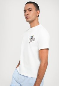 White cotton t-shirt with short sleeves, featuring a colorful graphic of a winged figure and a banner. Model wears striped shorts.