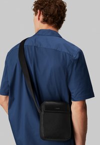 Borsa a tracolla nera in pelle testurizzata con design logo in rilievo; presenta un pattina superiore e una tracolla regolabile.