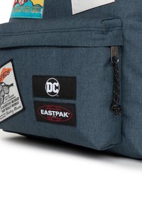 Tummansininen kangaskassi, jossa on useita merkkejä, mukaan lukien DC- ja Eastpak-logot, vetoketju ja kestävä tekstuuri.