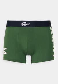 Gröna kalsonger med marinblå midjeband som har en vit krokodil-logotyp och "LACOSTE" tryckt i vitt längs sidan. Slät textur.