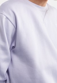 Osoba v svetle levandulovém svetru s kulatým výstřihem, na němž je viditelný V-šv detail u výstřihu.