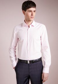 Camisa de vestir de manga larga color rosa claro con cuello, botonadura frontal y textura suave, combinada con pantalones oscuros de patrón y un cinturón negro.