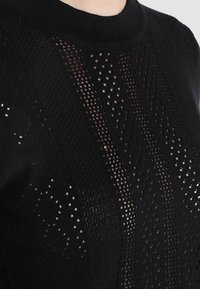 Černý pletený pulovr s vzorovaným designem, otevřenými průsvitnými částmi a vysokým límcem. Materiál je měkký s texturovaným povrchem.