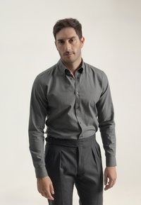 Camisa gris de botones y de manga larga, hecha de tela texturizada, combinada con pantalones oscuros de cintura alta y pliegues, con un cinturón en la cintura.