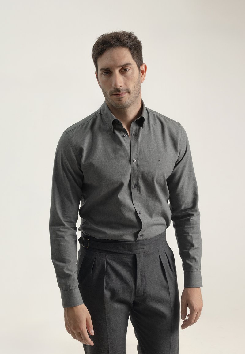 Camisa gris de botones y de manga larga, hecha de tela texturizada, combinada con pantalones oscuros de cintura alta y pliegues, con un cinturón en la cintura.