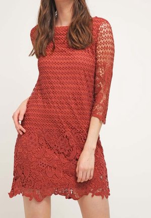 Kobieta w sięgającej do kolan sukience zrobionej na szydełku, w kolorze rdzawo-pomarańczowym, z wzorem kwiatowym i koronkowymi rękawami do łokcia, stojąca na jednolitym tle.