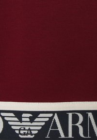 Burgundská mikina s texturovanou látkou a kontrastním bílým a tmavě modrým logem po obvodu, zobrazující emblém orla.