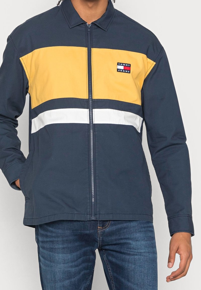 Marineblå och gul jacka med dragkedja och vit horisontell rand, har en krage och en liten logotyp på bröstet.