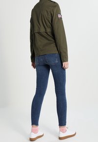 Oliwkowa kurtka wojskowa z naszywką, w zestawie z niebieskimi skinny jeansami i białymi trampkami z różowymi detalami skarpetek.