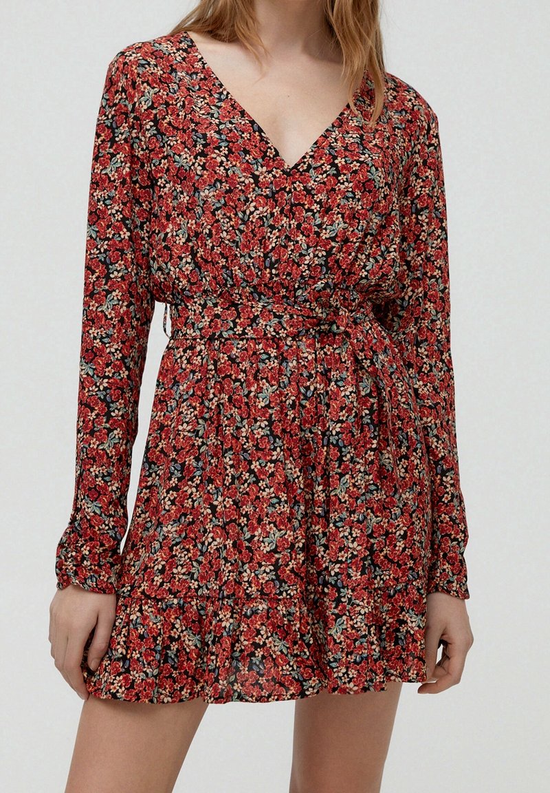 Femme portant une robe portefeuille rouge à fleurs à manches longues avec un décolleté en V et une ceinture à nouer à la taille.