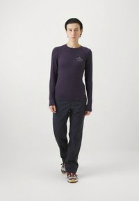 Long-sleeved purple shirt with a round neckline and a small logo on the left chest, paired with dark pants and sneakers.