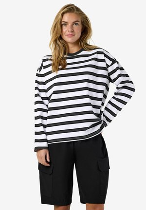 Młoda kobieta z długimi blond włosami, ubrana w biało-czarną poziomo prążkowaną bluzkę z długim rękawem oraz czarne spodnie cargo, stojąca z jedną ręką na biodrze.