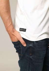 Hombre con camiseta blanca con etiqueta negra y jeans azul oscuro, con la mano izquierda parcialmente en el bolsillo delantero.