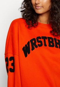 Sudadera naranja con texto bordado en negro que dice "WRSTBH" y el número "23" en la manga izquierda. Tejido suave, corte relajado.