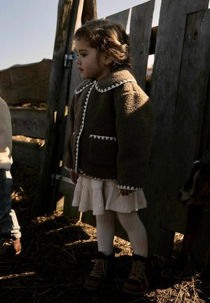 Ragazza giovane con giacca marrone calda e gonna bianca in piedi vicino a una recinzione di legno in un ambiente rustico all'aperto con un cane parzialmente visibile nelle vicinanze.