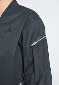 Melns bomber žakets ar rāvējslēdzēja piedurknes kabatu, izšūtu logo uz krūts un pogu aizdari apakšā.