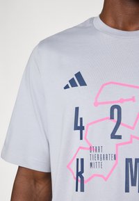T-shirt en coton gris clair avec un motif graphique géométrique bleu marine et rose vif, comportant des chiffres et le texte "START TIERGARTEN MITTE."