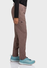 Braune Bergpants mit mehreren Reißverschlusstaschen, einer schmalen Passform und verstärkten Nähten. Kombiniert mit blauen Sportschuhen.