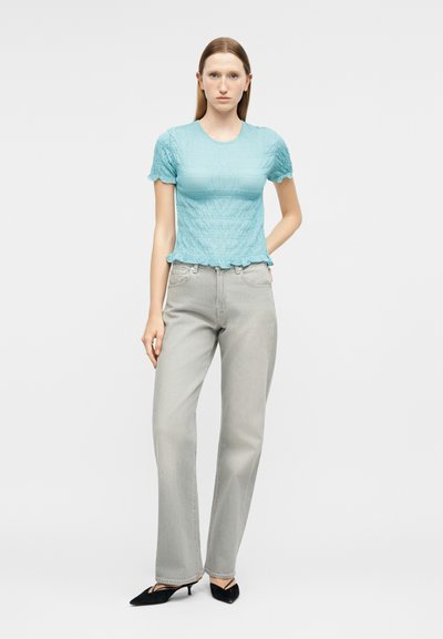 Γυναίκα που στέκεται με το χέρι στην τσέπη, φορώντας μια υφασμάτινη ανοιχτό μπλε μπλούζα με κοντά μανίκια, ανοιχτό γκρι τζιν με ίσια μπατζάκια και μαύρες γόβες με χαμηλό τακούνι.