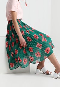Gonna plissettata floreale verde, caratterizzata da grandi fiori rossi; abbinata a una maglietta rosa chiaro e sandali bianchi con tacco a blocco.