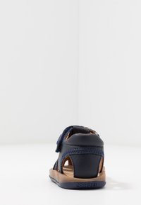Marinblå lädersandal med öppen design, justerbart rem, och brun gummisula. Har synlig sömnad och varumärkeslogo på sulan.