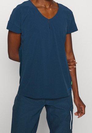 Personne portant un t-shirt bleu marine à manches courtes et col en V, avec un pantalon bleu marine assorti avec une poche latérale, debout avec une main sur son bras.