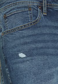 Jeans de mezclilla azul con un diseño estándar de cinco bolsillos, con una pequeña área desgastada en el muslo y costuras en amarillo contrastante.