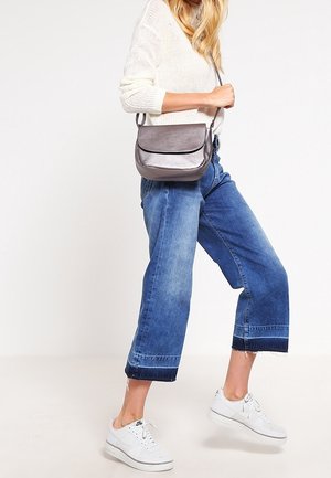 Grijze leren crossbodytas met een gebogen klep, gecombineerd met een lichtgeverfde trui en cropped blauwe jeans. Witte sneakers maken de outfit compleet.