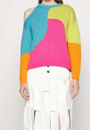 Vrouw draagt een meerkleurige trui met blauwe, roze, gele en oranje vlakken en een witte gelaagde rok met knoopdetails.