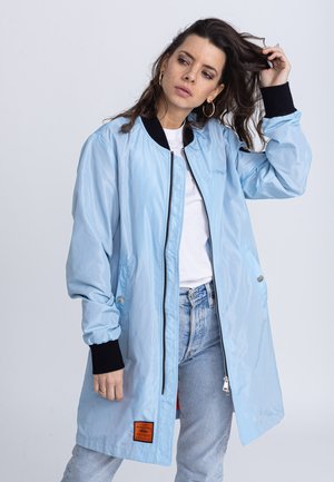 Lichtblauwe bomberjack met een ritssluiting aan de voorkant, zwarte ribgebreide manchetten, zijzakken en een oranje labeldetail aan de onderrand. Glad, glanzend materiaal.