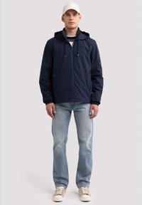 Tumesinine kapuutsiga jakk, millel on esiotsas lukku, reguleeritavad tõmblukkusid ja küljetaskud; koos helesiniste teksade ja beežide tossudega.