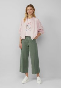 Veste bomber rose clair portée sur une chemise blanche à motifs floraux, associée à un pantalon large vert olive et des baskets blanches. Design simple, coupe décontractée.