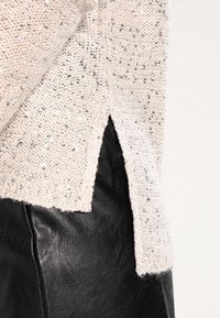 Suéter beige claro con textura moteada y lentejuelas, con una abertura lateral. Combinado con una falda de cuero negra, destacando materiales contrastantes.