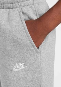 Šedé bavlnené teplákové nohavice so bočnou vreckom, s bielym vyšitým logom Nike na spodku. Hladká textúra a elastický pás.