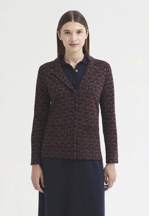 Femme aux cheveux bruns et raides portant une veste boutonnée bleu foncé à motifs sur une robe bleu marine, debout devant un fond blanc uni.