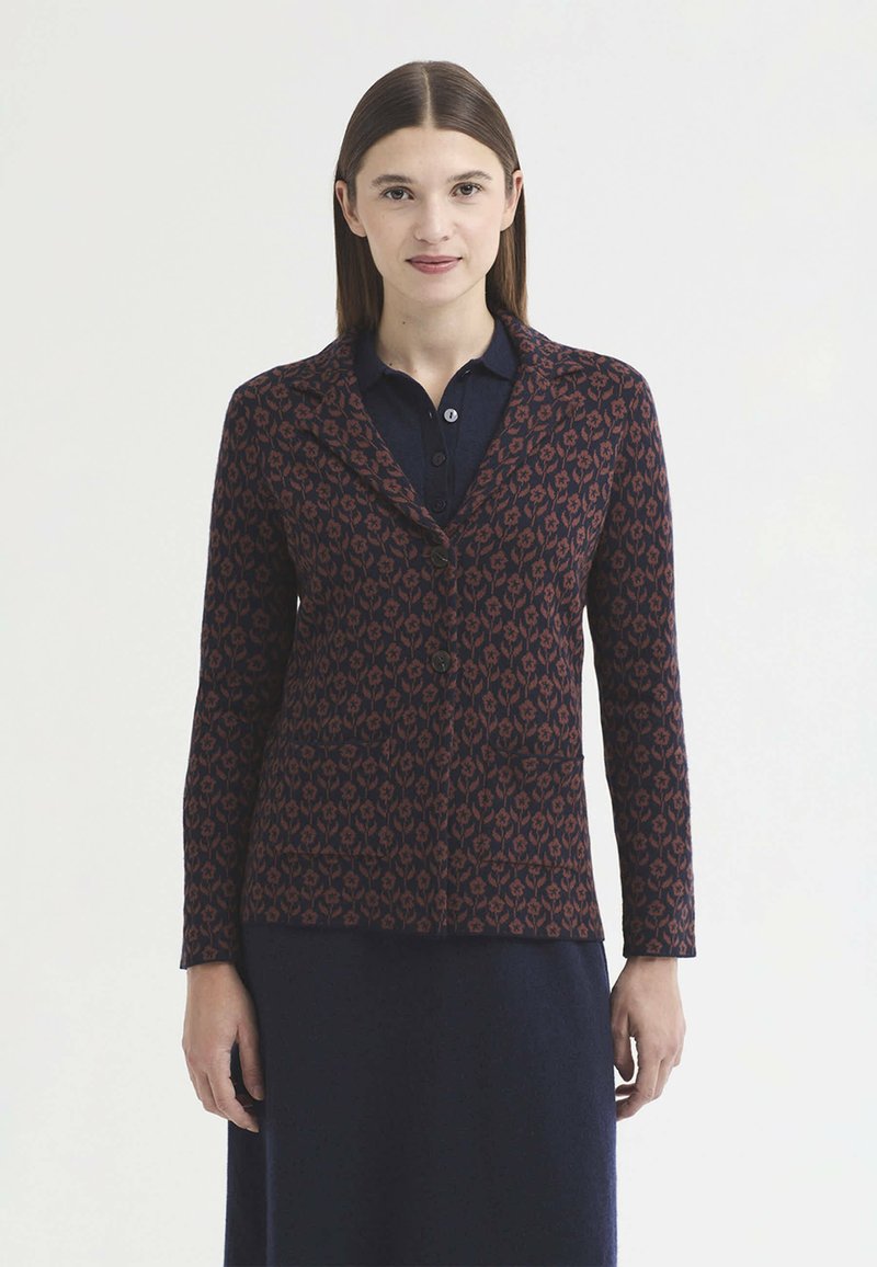 Femme aux cheveux bruns et raides portant une veste boutonnée bleu foncé à motifs sur une robe bleu marine, debout devant un fond blanc uni.