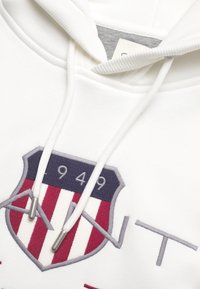 Weiße Kapuzenjacke mit Kordelzug und gerippten Akzenten. Verfügt über ein gesticktes Wappenlogo in Marineblau, Rot und Grau mit "949"-Detail.