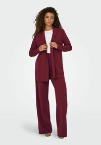 Costume bordeaux composé d'un long blazer et de pantalons à jambes larges, associé à un haut blanc. Tissu lisse avec une coupe ajustée.