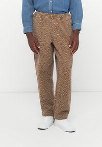 Brown patterned pants with an elastic waistband, featuring a textured fabric and gray circular designs, paired with white sneakers.