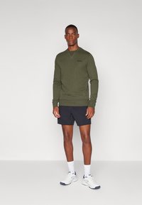 Camisa de manga larga verde oliva con cuello redondo y logotipo, combinada con pantalones cortos negros y zapatillas deportivas blancas. Diseño sencillo, ajuste casual.