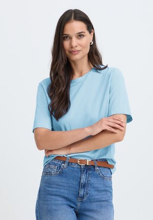 Femme aux longs cheveux foncés portant un t-shirt bleu clair, un jean bleu et une ceinture marron, debout les bras croisés devant un fond uni.