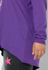 Lilla langærmet skjorte med afrundet kant og struktureret overflade, parret med sorte leggings med et pink stjernedesign på låret.