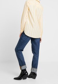 Camisa de rayas amarillas y blancas de manga larga, jeans azul con puños doblados, y botas negras de tacón con un acabado brillante, vistas desde un lado.