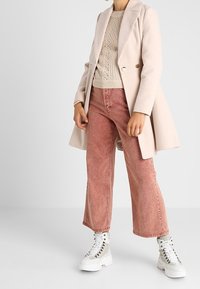 Světle růžový kabát, krémový pletený svetr, vybledlé růžové široké kalhoty a bílé tenisky s vysokým střihem a stříbrnými akcenty. Minimalistický design.