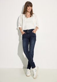 Jean skinny bleu foncé avec une taille à double bouton, associé à un débardeur blanc et une chemise en lin légère, complété par des baskets blanches.
