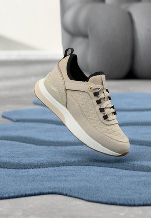 Bež superga z reliefnimi paneli in črnim okrasjem, ki visi nad valovito modro preprogo na sivem tlaku z zamegljeno skulpturo v ozadju.