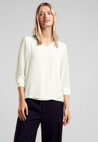 Blusa bianca leggera con scollatura a V e maniche a tre quarti. Presenta una texture liscia e una vestibilità comoda, abbinata a pantaloni neri.