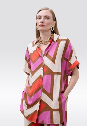 Mujer con camisa oversize con patrón geométrico en rosa, marrón, rojo y crema, con un gran collar de cadena dorada y pendientes a juego, manos en los bolsillos.