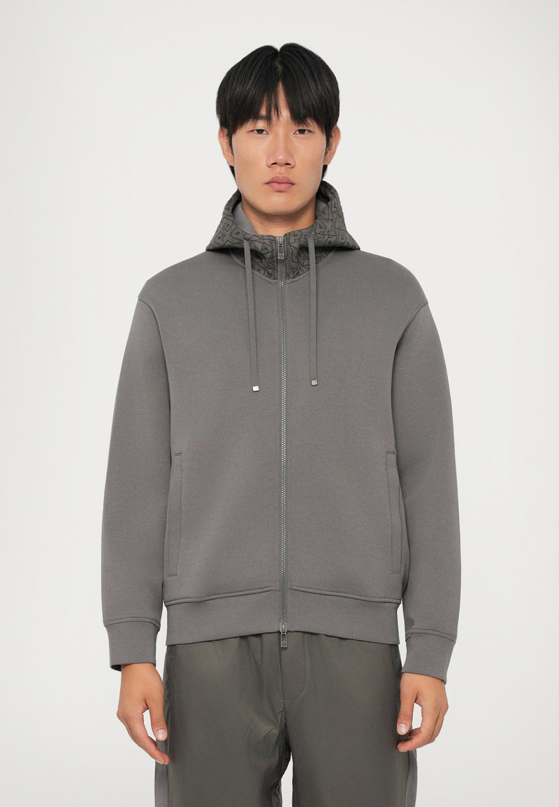Felpa grigia con zip, cappuccio con motivo, lacci, tasche laterali e polsini a costine. Il materiale sembra morbido e testurizzato. Design minimalista.