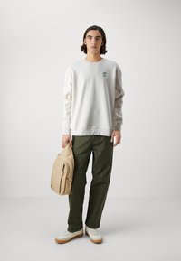Biała bluza z zielonym logo, oliwkowe spodnie, beżowy plecak i białe sneakersy z gumową podeszwą. Prosta, casualowa stylizacja.
